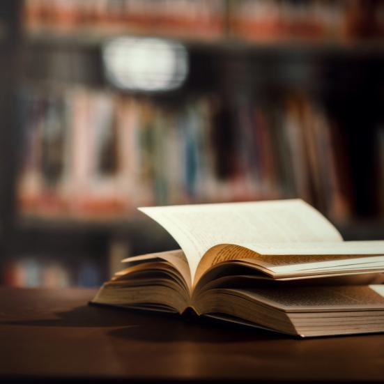 Åben bog med blafrende sider, der ligger på ryggen på et bord. I baggrunden ses reoler med bøger i.