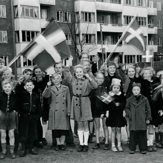 Foto i sort/hvid af en masse børn, der smiler og vifter med dannebrogsflag foran en lejlighedsbygning.