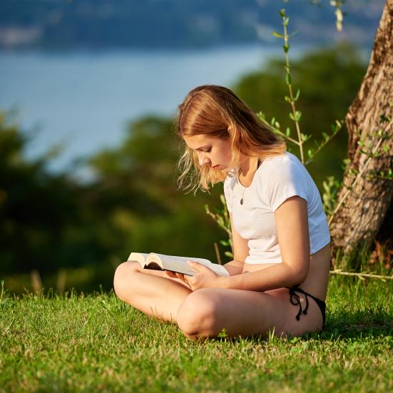 Kvinde, der sidder på græsset og læser en bog med natur i baggrunden.