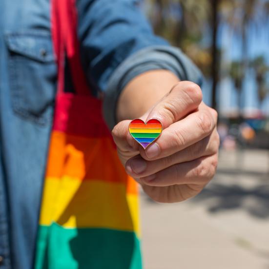 Person, der holder en hjerteformet og regnbuefarvet pin frem mod kameraet.